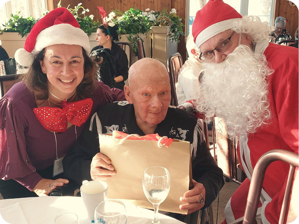 Trois personnes, dont un père Noel qui regardent la caméra en souriant avec un cadeau à la main.